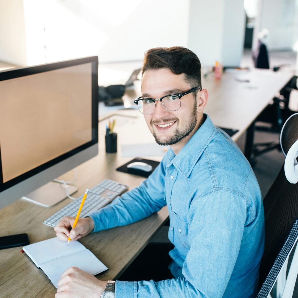 Young man in glassess is working at his workplace in office. He wears blue shirt.  He is writing in notebook and smiling to the camera.